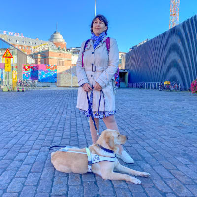 En kvinna står på Hagnästorget med sin ledarhund. 