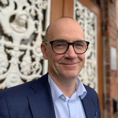 Mansperson klädd i blå kostym ler och ser in i kameran. I bakgrunden husfasad och port.