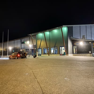 En grå hallbyggnad, kväll, byggnaden är upplyst. Bilar står parkerade framför byggnaden, barn på väg in i hallen. Raseborgs ishall.