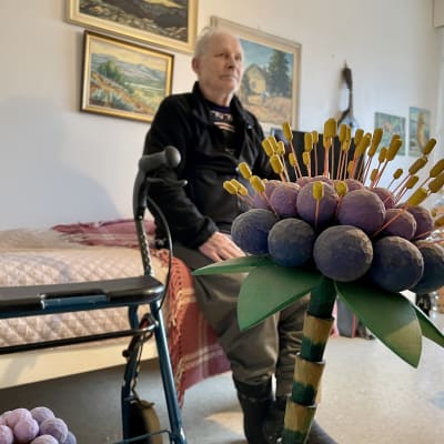 Puinen veistos huoneen lattialla, taustalla istuu vanha mies sängyn reunalla.