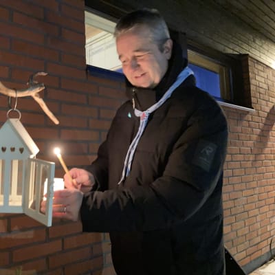 Mies sytyttää tuohuksen lyhdyssä palavalla kynttilällä ulkona talon edustalla.