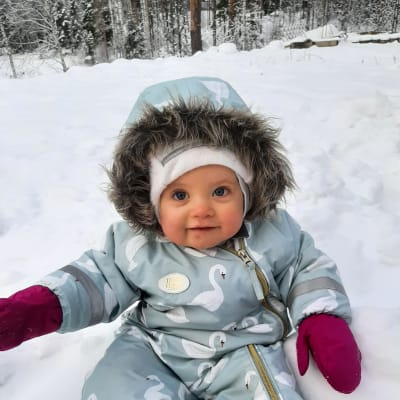 En bebis i blå vinteroverall sitter i en snödriva och ler.