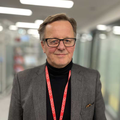En man med glasögon och kavaj ser glad ut och tittar rakt in i kameran, där han står i en korridor. 