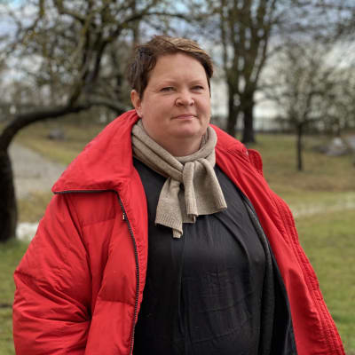 En kvinna med kort hår i röd rock framför några träd.