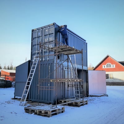 Två kontainrar staplade på varandra. Person som bygger.