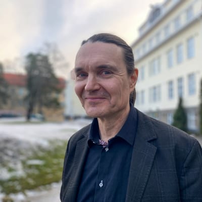 Personporträtt på man som ler och tittar in i kameran. Han är klädd är mörkblå skjorta och kavaj, i bakgrunden våningshus med ljusgul rappning.