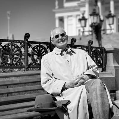 Svartvit bild på Matti Klinge som sitter på en bänk på torget framför domkyrkan i Helsingfors, leende och klädd i solglasögon. 