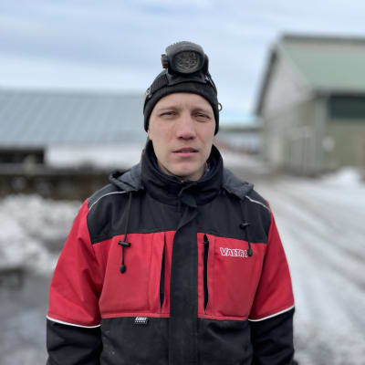 En man i arbetsoverall står ute på en pälsfarm med mössa och pannlampa i snömodden. 
