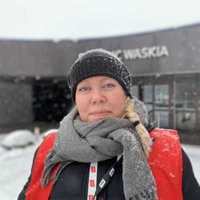 Kvinna i mössa, halsduk och ytterrock med röd väst står utomhus i snöfall. Hon ler och ser in i kameran, i bakgrunden hotellentré.