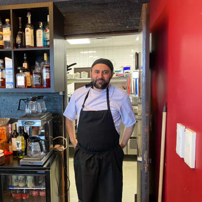 En man i kockkläder står i dörröppningen till ett kök i en restaurang. Han tittar mot kameran och ler.