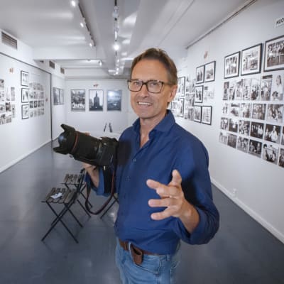 En man håller en systemkamera i handen, i bakgrunden väggar täckta med fotografier