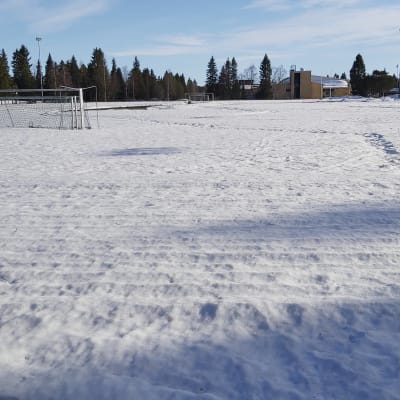 Kuvassa jalkapallokenttä kauttaaltaan lumenpeitossa. Keskellä kenttää jalkapallomaali, taustalla koulurakennus.