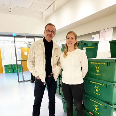 Två personer står i en korridor där det finns staplar med flyttlådor längs väggarna. De ser in i kameran och ler.