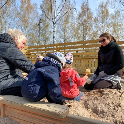 Två kvinnor sitter vid en sandlåda tillsammans med flera barn. En av kvinnorna ler.