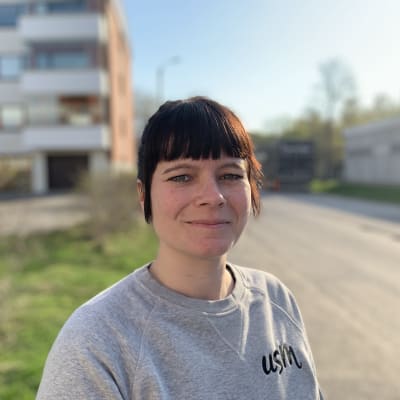 Porträtt på kvinna med mörkt hår och ljusgrå blus. Hon ler svagt och tittar in i kameran. I bakgrunden bostadshus och gröna gräsmattor.