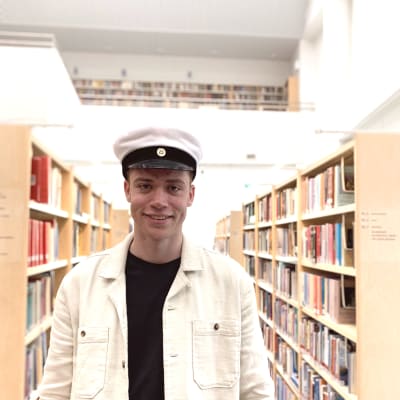 Personporträtt på ung man, han har studentmössa på huvudet, bär ljus jacka och ler. I bakgrunden bokhyllor i biblioteksmiljö.