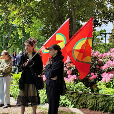 Två kvinnor håller i PKK-flaggor på en demonstration utomhus vid en park.