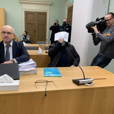 Två män sitter i en domstol bakom ett bord, till vänster advokaten med glasögon och till höger en man som gömmer sitt ansikte bakom ett vitt papper.