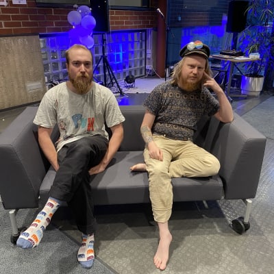 Taideakatemian opiskelijat Erno Beuker ja Tuomas Vainionpää istuvat mustalla sohvalla. Taustalla näkyy punainen tiiliseinä ja lasitiiliä. Sohvan takana on kaiuttimia. Beuker on pukeunut harmaaseen t-paitaan ja mustiin housuihin. Hänellä on jalassaan värikkäät sukat. Vainionpäällä on päässään musta hattu, tumma, kuviollinen t-paita ja ruskeat housut. Hänellä ei ole sukkia.