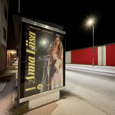 Anna Liisa -teatteriesityksen juliste linja-autopysäkillä. Tiellä on lunta.