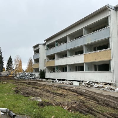 Purettava kerrostalo, purkujätettä maassa ja työkoneet käsittelemässä purkujätettä taustalla.