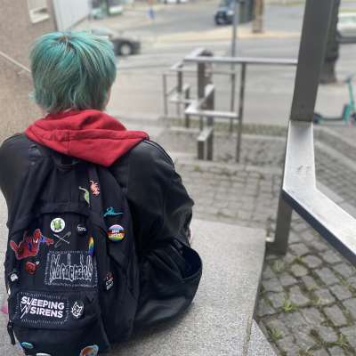 En ung person sitter på en trappa. Han har blått hår och en ryggsäck på ryggen med regnbågssymboler. Bilden är tagen bakifrån.