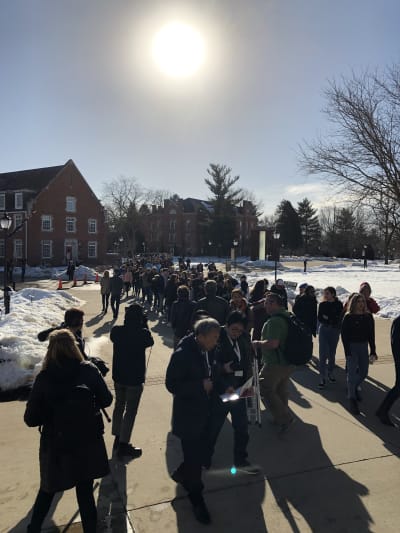Flera människor köar till Elizabeth Warrens evenemang. Solen skiner och snön ligger i drivor vid vägen som annars är bar.
