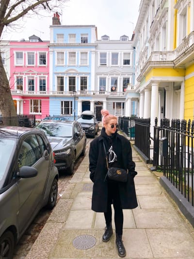 Nuori nainen tukka sykeröllä aurinkolaseissaan astelee jalkakäytävällä Lontoossa, taustalla värikkäitä taloja.