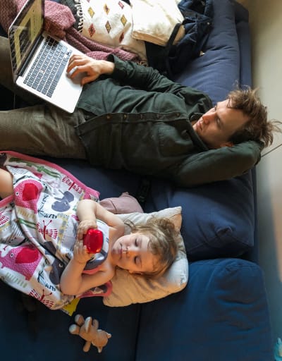Pappa och dotter ligger i soffan bredvid varandra. Pappa har ena handen på sin bärbara dator och andra handen under huvudet, medan dottern ligger under ett täcket och knapprar på en leksak.