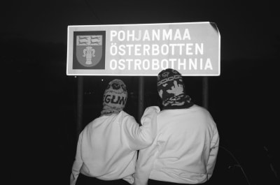 Två rappare i skidluvor står vända mot en österbotten-skylt.