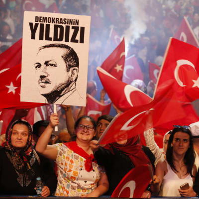 En folkmassa viftar med Turkiets röda flagga. En person håller upp president Erdoğans porträtt med texten "Demokratins stjärna".