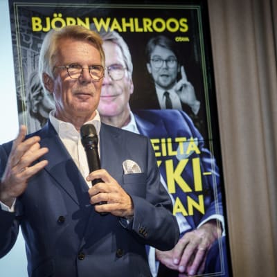 Björn Wahlroos lavalla puhumassa mikrofoniin. Taustana kuva hänen kirjansa kannesta.