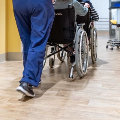 Vårdare skuffar en äldre person i rullstol.