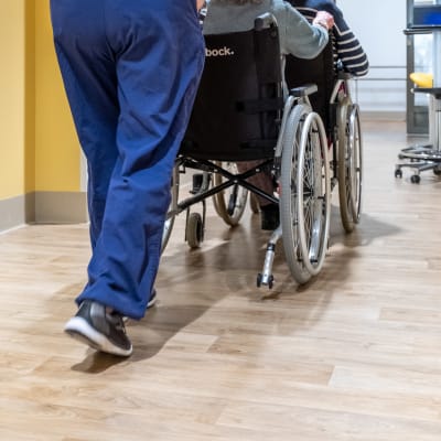 Vårdare skuffar en äldre person i rullstol.