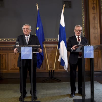 Jean-Claude Juncker och Antti Rinne står brevid varandra i varsin talarstol. Bakom dem syns både Finlands och EUs flagga