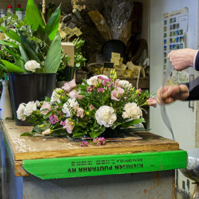 En blomsteruppsättning för en begravning ligger på ett bord i en blomsterhandel. Till höger syns en i personalen som håller på och gör en rosett.