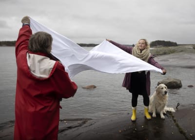 Näyttelijä Nelly Juulia Kärkkäinen ja äitinsä Gitta Oksanen hulmuttavat valkoista lakanaa rantakalliolla, vieressä koira. Promokuva Kansallisteatterin Pudota-esityksestä, joka kertoo tahattomasta lapsettomuudesta.