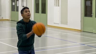 En ung pojke, cirka fjorton år, står i beråd att kasta en basketboll i en korg i en gymnastiksal.