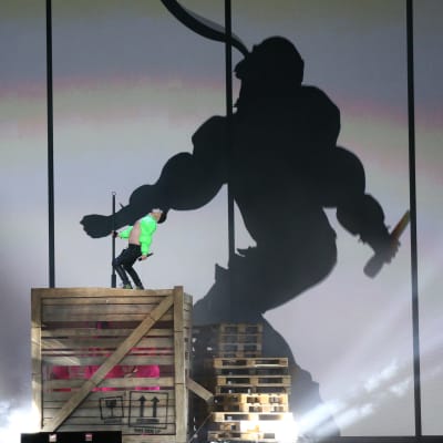 En man iklädd neongrön bolero står på en stor trälåda och dansar. I bakgrunden syns hans skugga projicerad på en stor skärm.