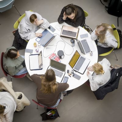 Studerande sitter runt runda bord. Alla har datorer framför sig på bordet där de jobbar.