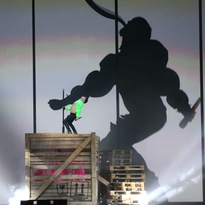 En man iklädd neongrön bolero står på en stor trälåda och dansar. I bakgrunden syns hans skugga projicerad på en stor skärm.