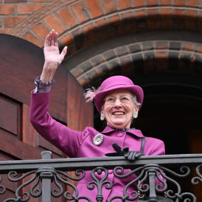 Drottning Margrethe iklädd lila hatt och kappa vinkar glatt på en balkong.