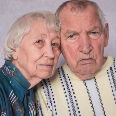 Ett äldre par i blå och gul blus. Det har lutat sina huvuden mot varaandra och ser ledsna eller bekymrade ut i sina ansikten