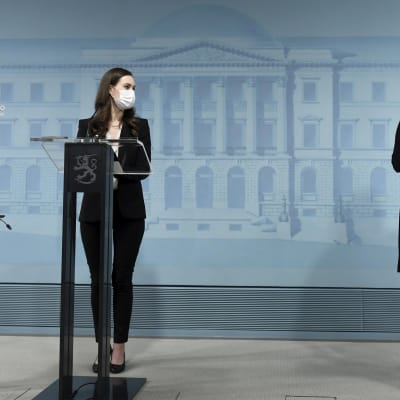 Statsminister Sanna Marin (till vänster) och arbetsminister Tuula Haatainen vid regeringens presskonferens. Båda har munskydd på sig och de står vid varsitt talarpodium.