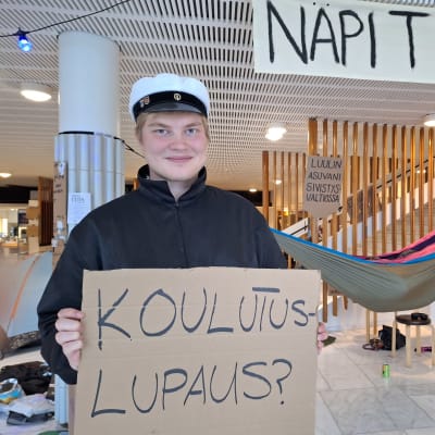 Ylioppilaslakkipäinen opiskelijapoika pitää yliopiston aulassa kädessään kylttiä jossa kysytään koulutuslupauksesta.