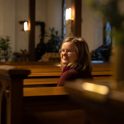 Elena Alakomi istuu kirkon penkissä ja katsoo kohti kameraa.