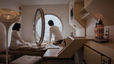Ett ultramodernt hotellrum där en man och kvinna sitter på sängen och ser ut genom ett runt fönster.