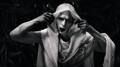 Svartvit bild på en blek Gorr the God Butcher (Christian Bale) som lyfter på en vit huvudduk och avslöjar sitt ansikte.