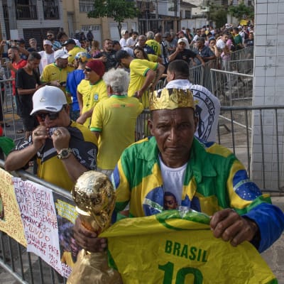 Fans i kö för att ta farväl av fotbollsspelaren Pelé. I förgrunden står en man iklädd en guldkrona och Brasiliens färger. I handen håller hans en kopia av världsmästerkapspokalen.