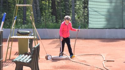 En kvinna på tennisplan. Hon spelar inte utan har just vattnat planen. Vattenslangen ligger på marken.